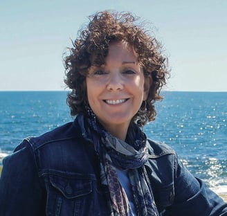 Aggie, with short curly hair and a denim jacket, stands in front of a bright blue sea.