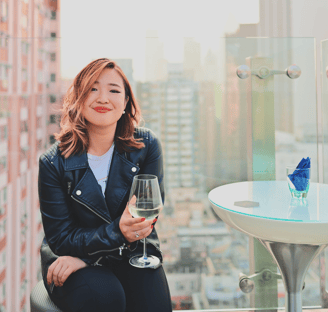 Jemimah Wei holds a glass of white wine and sits, smiling, on a rooftop overlooking a cityscape.