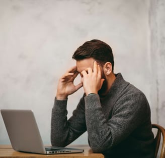 a man sitting at a desk with a laptop computer. holding his head with his hands