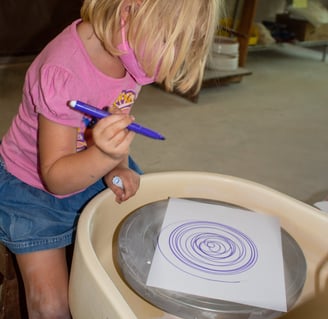 Young girl sitting at a potters wheel holding a marker, making spin art.