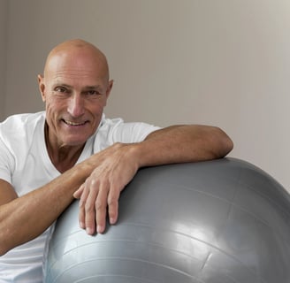 Homem careca sentado no chao ao lado de uma bola de pilates