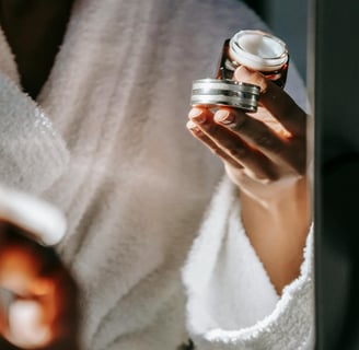A woman looking at a mirror, wearing an eye cream with one hand and holding the cream container on the other hand.