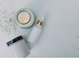 Image showing from above a white candle, a glass container of a moisturizer and a serum, all placed on a white marble surface