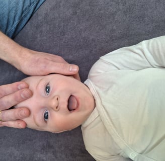 een baby ligt op zijn rug en lacht terwijl 2handen zijn hoofd vasthouden.
