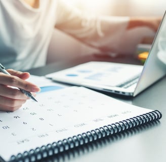 someone working at a desk with a calendar, laptop, and pen in hand