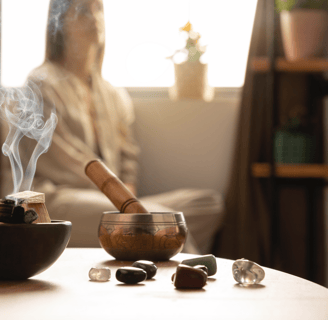 Table with crystals, palo santo, tibetan bowl, and a woman meditating in the background