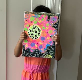 little girl with dark hair in a pink and orange dress holds colorful painting in front of her face