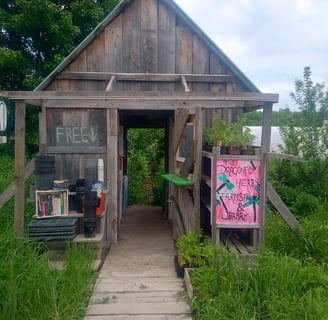A wooden farmstand with an open entrance, featuring signs for organic eggs, free produce, and 'The D