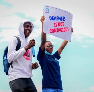 A man in a white hoodie, standing next to a woman who is holding up a banner that says "Deafness is not contagious"