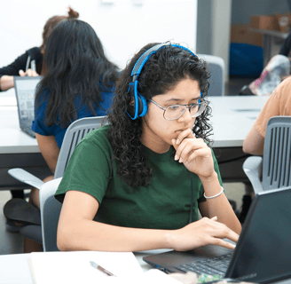 Mulher jovem de óculos e fones de ouvidos, utilizando notebook sobre a mesa, em uma sala de aula
