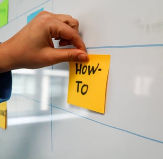 A hand posting a sticky note with the phrase "how-to" written on it.