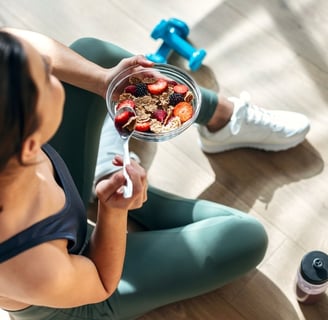 A woman eating cereal and fruits from a bowl, with small weights and a bottle of water on the floor of the gym studio