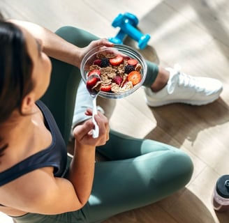 A woman in gym clothes sitting on the floor of a studio with a pair of weights, eating a healthy bowl of fruit and cereal