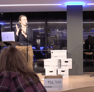 A white woman speaks at a microphone beside a pile of white fileboxes