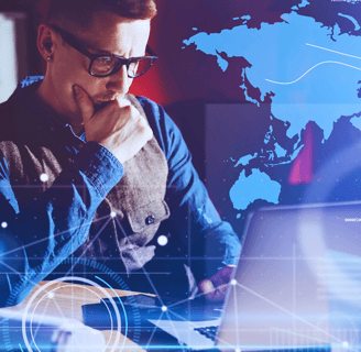 Business leader in glasses works on a laptop at a desk, one hand over his mouth, with a holographic continent behind him.