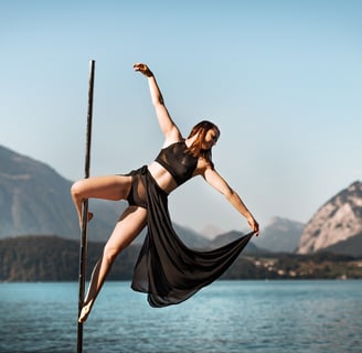 Sabrina Bel Pole doing a cupid pole trick outdoors, with a lake and mountains in the background.