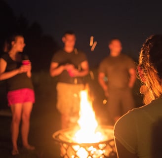 A nighttime gathering around a glowing campfire, with silhouettes of people standing in a circle. The warm firelight illumina