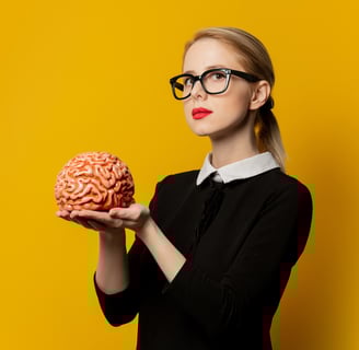 a blond woman with black glasses, holding a brain. she's on a yellow background.