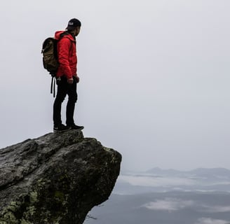 A man in a red jacket standing on a cliff rock, looking out at the plains against a cloudy grey sky