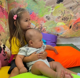 Niña abrazando bebe sentados entre cojines de colores vibrantes en Ukelele Jardín Infantil