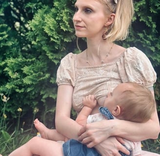 Mother sitting in front of a green bush, wearing a bow in her hair, holding and nursing her baby & looking away peacefully