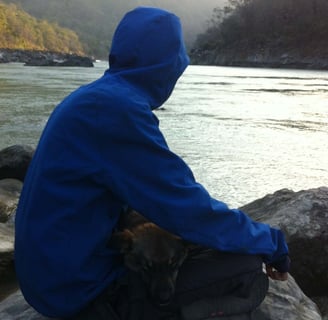 a man in a blue jacket sitting on a rock by the side o river ganga with dog sleeping in his lap