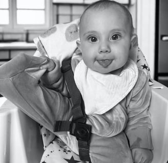 Black and white photo of smiling baby in high chair with tongue out, showing natural baby joy, innocence, and purity.