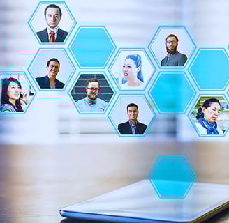 A tablet rests on a desk as holographic honeycomb shapes with headshots of businesspeople emerge from the screen.