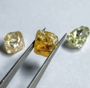 The image features three rough, uncut diamonds resting on a smooth, light-colored surface. A pair of