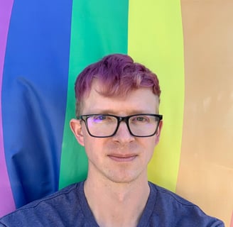 Portrait of Tomkins with purple hair with a rainbow flag as a backdrop.