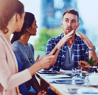 A man sits at a table, using hand gestures to explain something to four others seated around him.