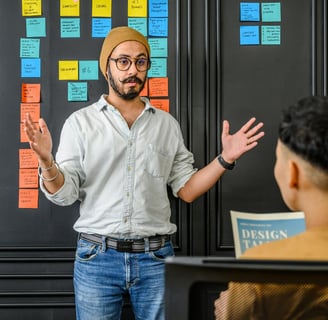 A casualy dressed man giving a talk standing infront of a board with colorful sticker notes on it