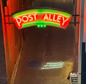 A neon sign saying post Allie with a holographic projection of Seattle #selfie museum underneath