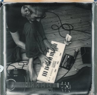 un musicien joue du clavier à même le sol, entouré d'une guitare, d'un micro et de câbles