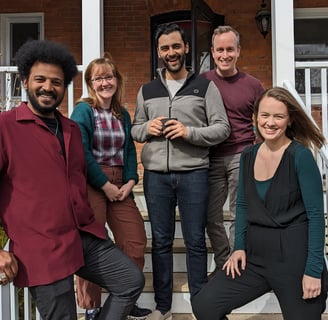 A group of five diverse artists pose on the steps of a brick house, smiling at the camera