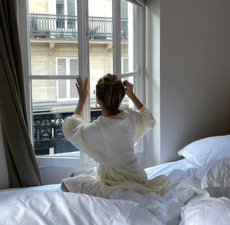 Woman in a robe sitting on a bed and tying her hair in front of a Parisian window — soft morning 