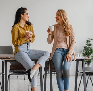 Two women talking and having coffee at a desk in an office, with a laptop on the table