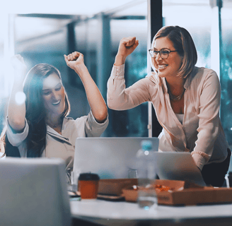 Group of leaders collaborates at a boardroom table, focusing on two female leaders celebrating their win with fist pumps