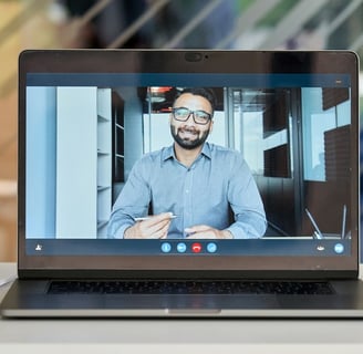 A laptop screen showing a video call with a man in a business shirt holding a pen