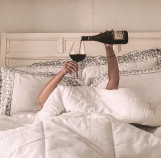 Woman lounging in bed pouring red wine — cozy, indulgent slow living moment