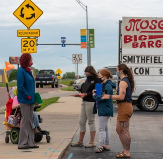 Felecia on a sidewalk next to a busy road speaking to a group of people with clipboards