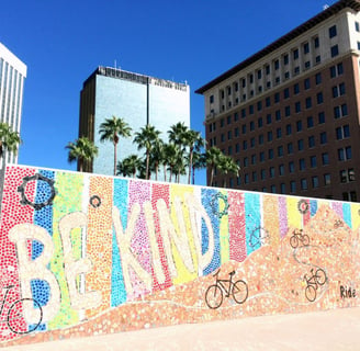 Big city picture with palm trees, tall buildings, and a mural spelling out "Be Kind" with bikes.