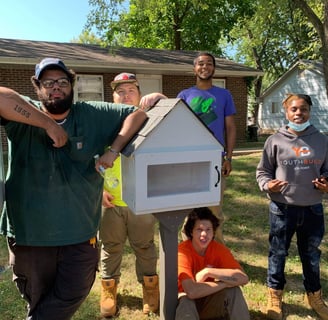 A group of kids and adults with Matthia, posing with a little library in front of a house