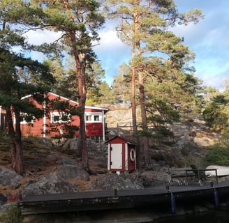 une maison rouge suédoise est au bord de l'eau , il y a des arbres autour de la maison.