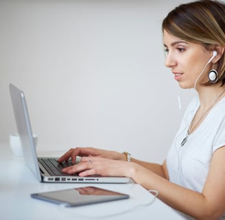 a woman with headphones, in a white shirt is sitting at a desk, typing