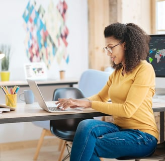 a woman wearing a mustard sweater and jeans, sitting at a desk working on a laptop computer