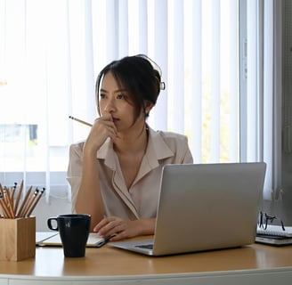 A thoughtful businesswoman in her office, sitting at her desk with an open laptop computer.