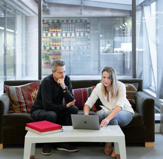 A man and woman sitting on a sofa in an office, looking at a Macbook.