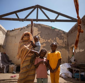 Sudanese woman and children standing inside a roofless home damaged by conflict.