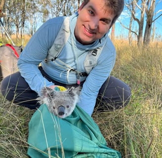 Ted Pavlic in hiking clothes holding a bag with a small koala head poking out of the bag. Eucalyptus trees in background.